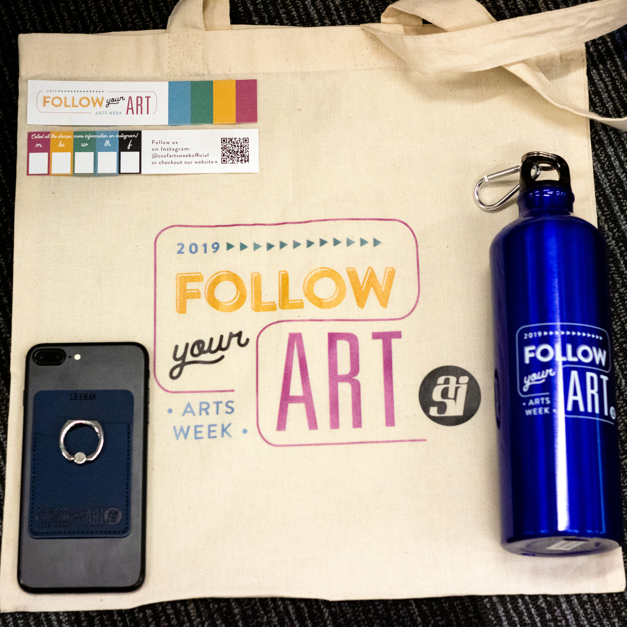 Arts Week 2019 canvas bag on the ground with bookmarks, cell phone back holder, and blue metal water bottle.