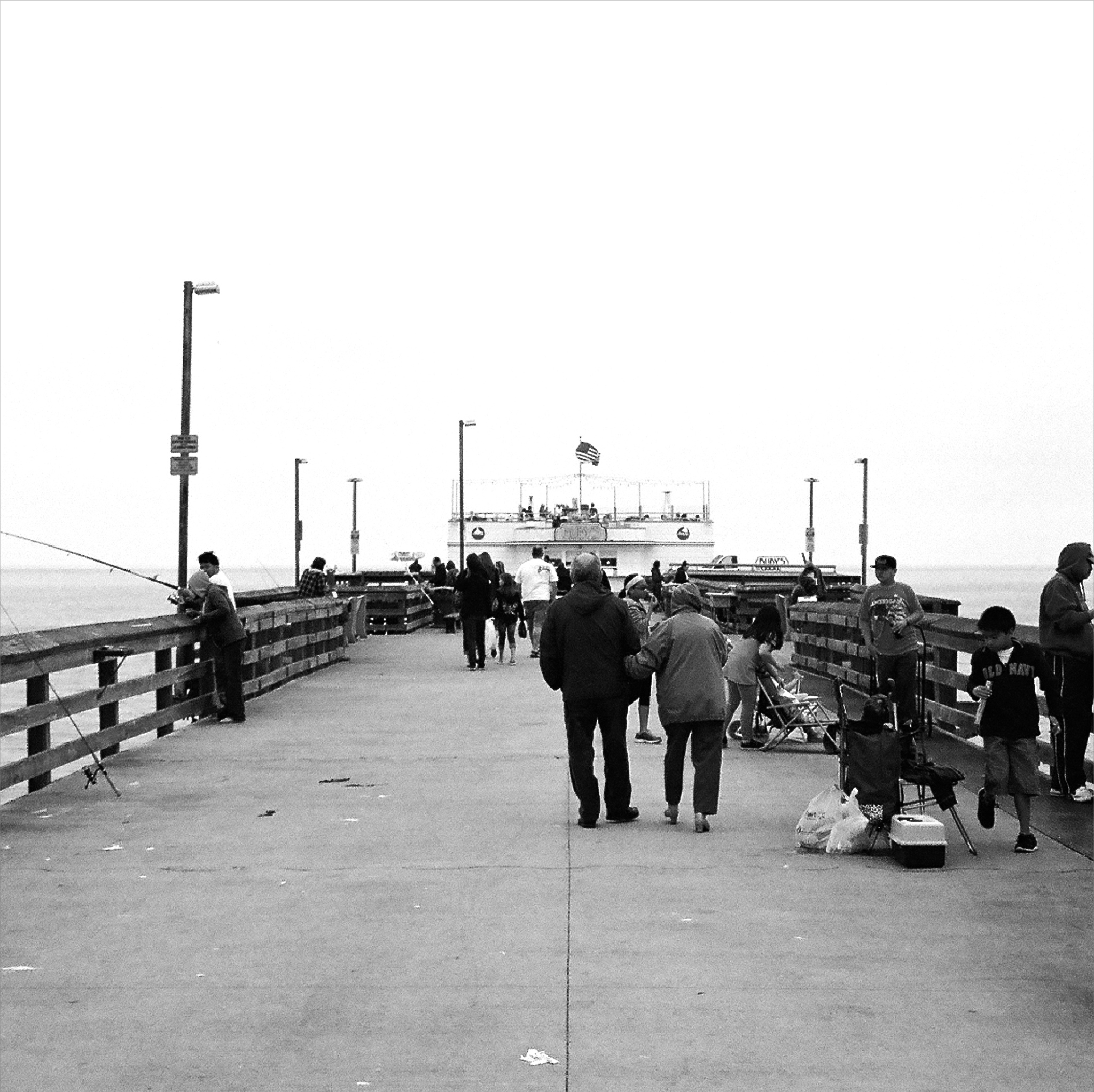 Square black and white photo of Balboa Pier with an elderly couple walking flanked with fishermen tending to their fishing rods.