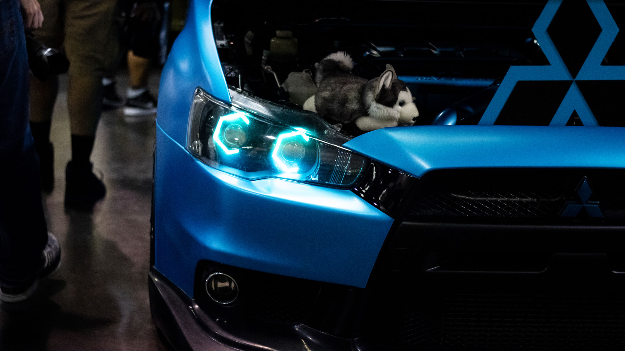 Front of a wrapped blue Mitsubishi with modified blue headlights and a wolf plushie on top of the car at SPOCOM Anaheim 2019.