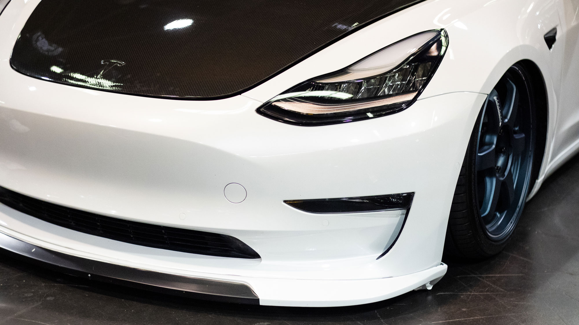 Photo of the front left side of a modified slammed/lowered white Tesla with a carbon fiber hood at SPOCOM Anaheim 2019.